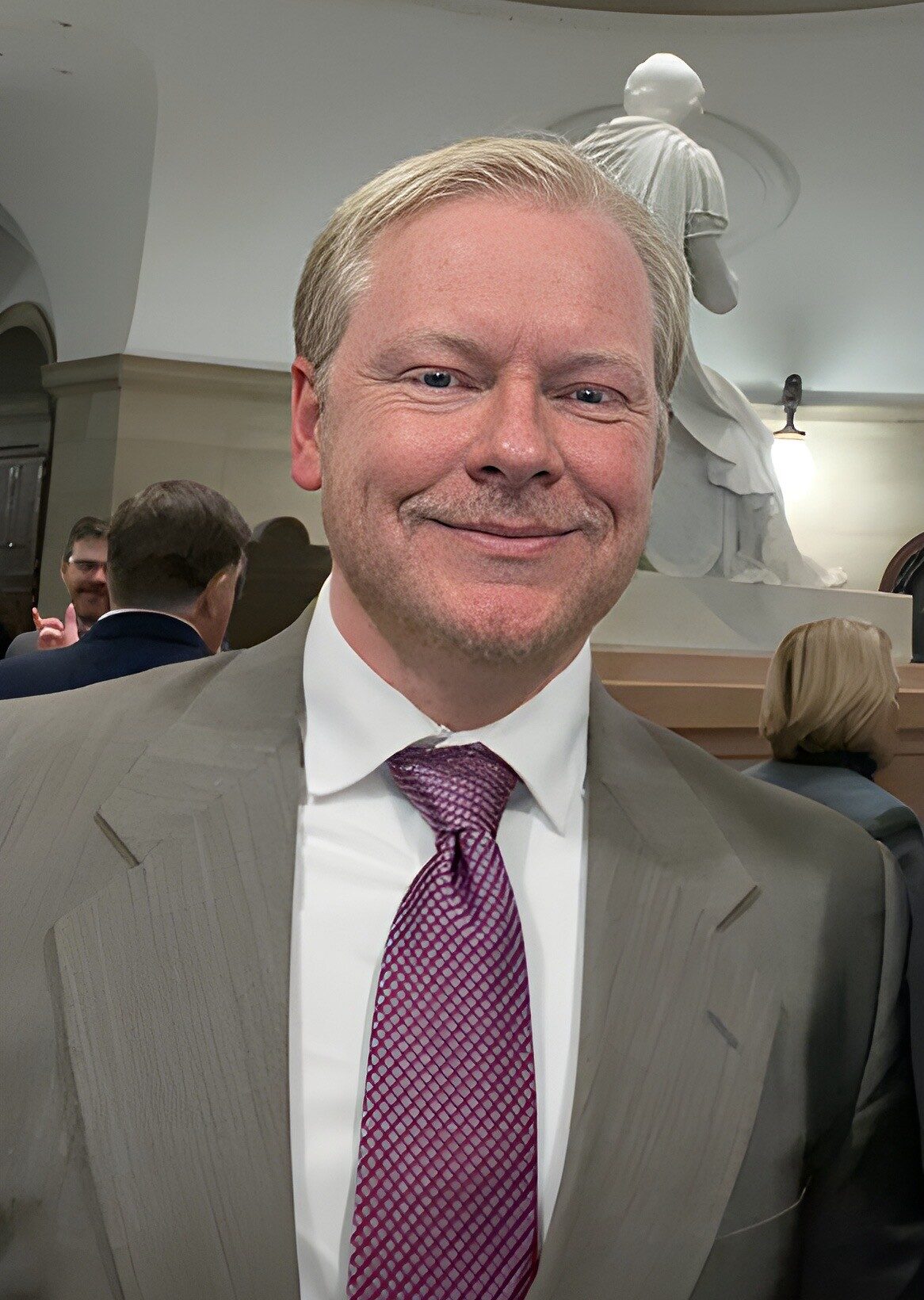 Cadman Kiker wearing suit and purple tie at indoor event with statue in background