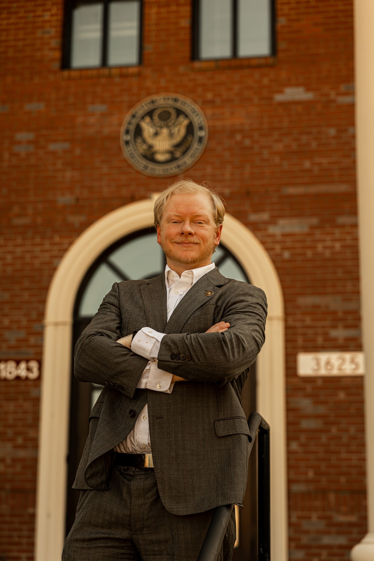 Cadman Kiker standing with arms crossed in gray suit in front of federal building with US seal