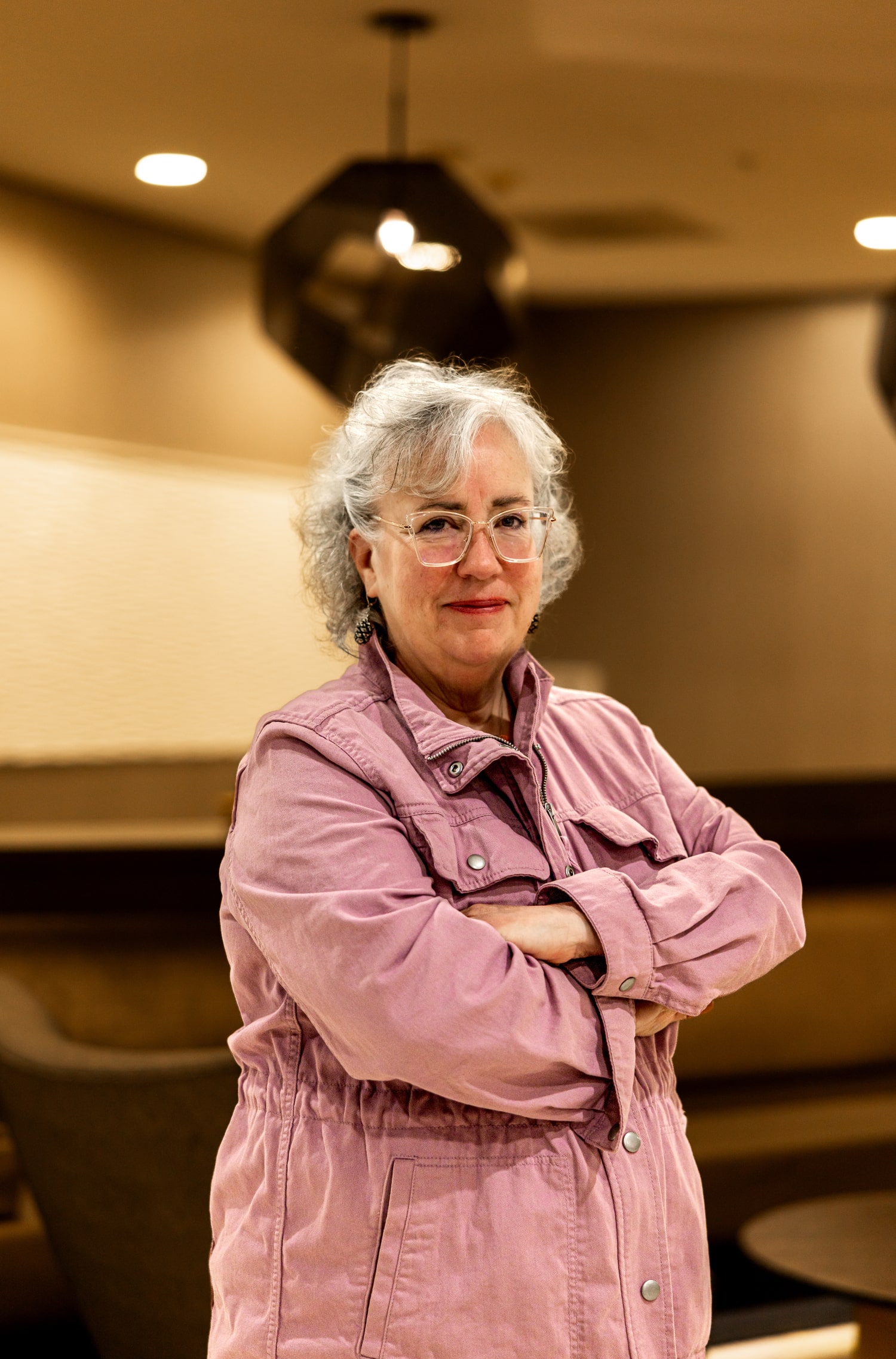 Genie Hughes posing with arms crossed in a pink jacket inside a lounge