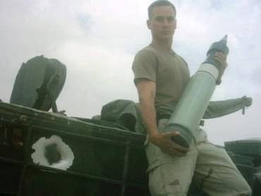 John Caulwell holding a large artillery round while standing on military equipment