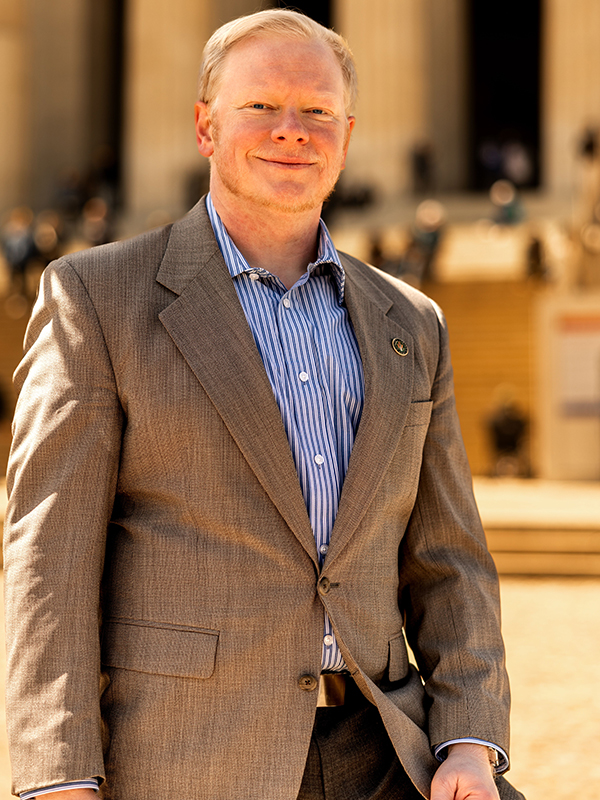 Cadman R. Kiker Military attorney Cadman Kiker standing outdoors, wearing striped shirt and tan suit jacket with building in background.