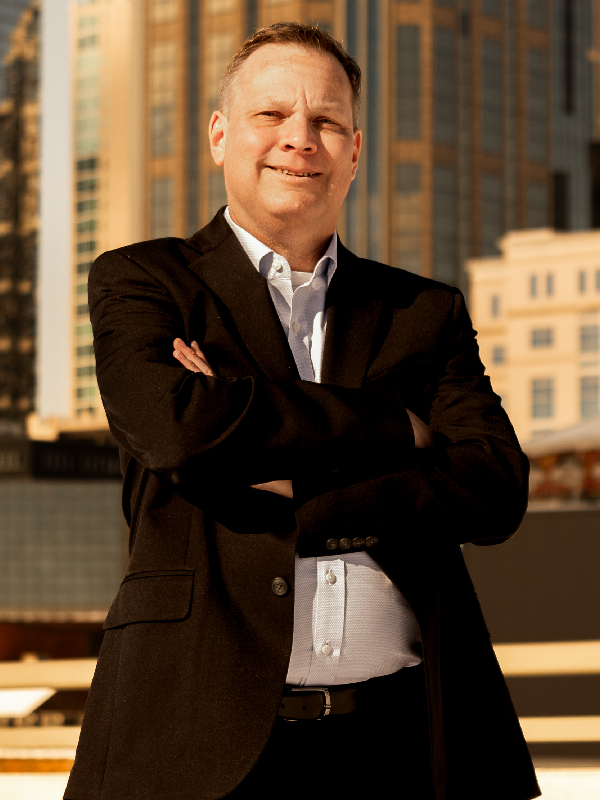 EJ Gladding Military attorney EJ Gladding standing outdoors with arms crossed, wearing black suit and light blue shirt, city buildings in background