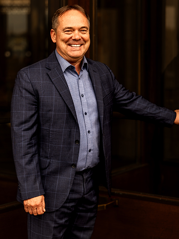 Scott Brokaw Military attorney Scott Brokaw standing indoors, wearing blue plaid suit and blue shirt, smiling