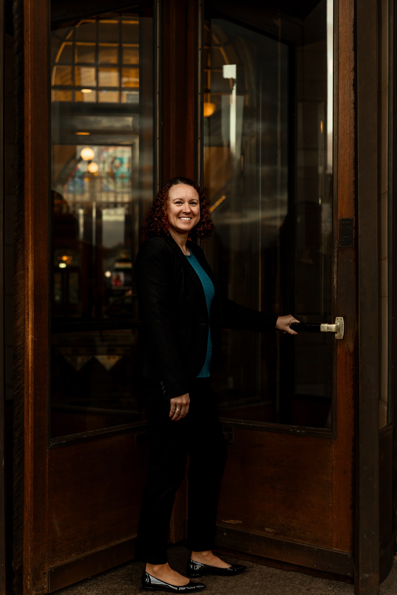 Victoria Camire standing by a large wooden doorway with a smile, holding the door handle