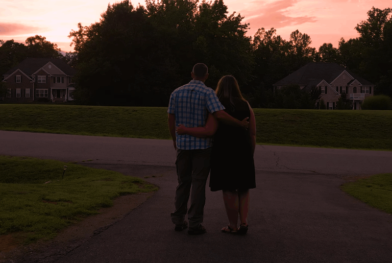 Jason and Audrey Byers standing arm in arm on a suburban street at sunset, looking toward houses and a glowing pink sky