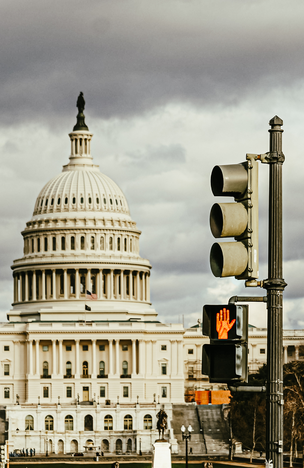 View of the U.S. Capitol dome with a traffic light and red pedestrian stop signal in the foreground under cloudy skies