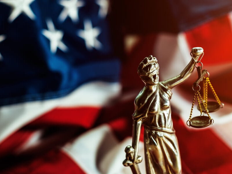 Bronze Lady Justice statue holding balanced scales in her raised right hand, with a blurred American flag behind her