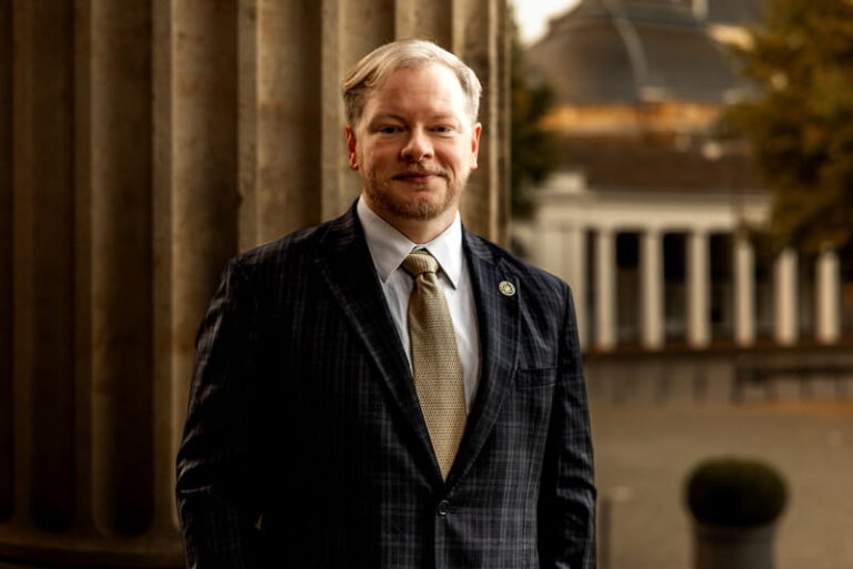 Cadman R. Kiker, III, military defense attorney in a dark plaid suit and gold tie, standing before stone columns