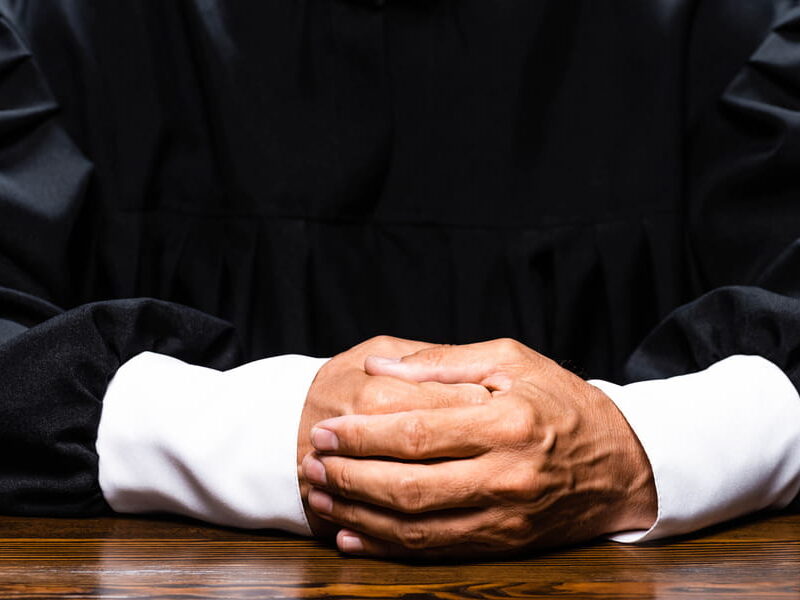 Close-up of a judge in a black robe with white shirt cuffs, hands folded together on a polished dark wood bench, face not visible
