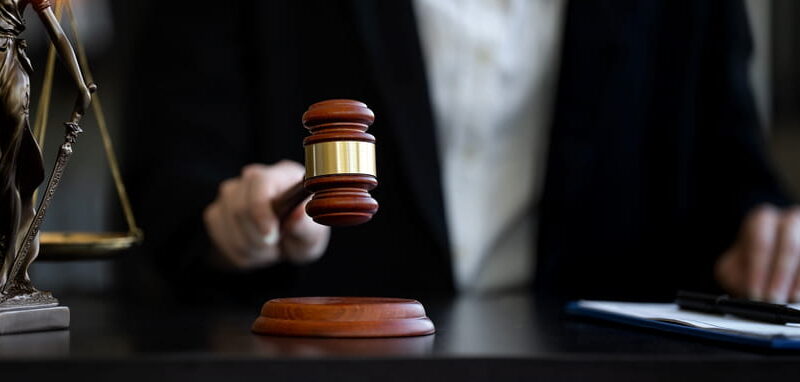 A judge holds a wooden gavel at a desk beside a bronze Lady Justice statue and a clipboard with a pen.