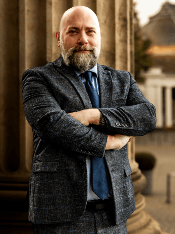 Matthew Krause in a checkered suit and blue tie, standing with arms crossed by stone columns outdoors