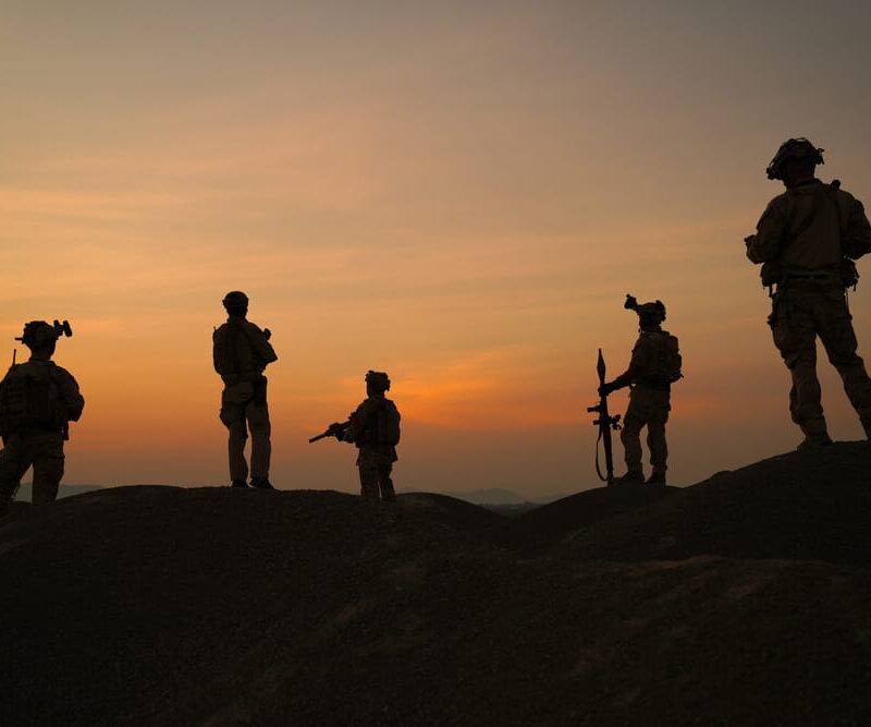 Silhouettes of five soldiers with rifles standing on a hilltop against an orange sunset sky