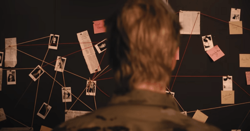 A soldier in camouflage uniform viewed from behind studying a wall of evidence with photos, news clippings, and notes connected by red string