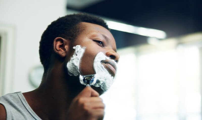 Young man in a grey tank top shaving his cheek with a cartridge razor, face covered in white shaving cream