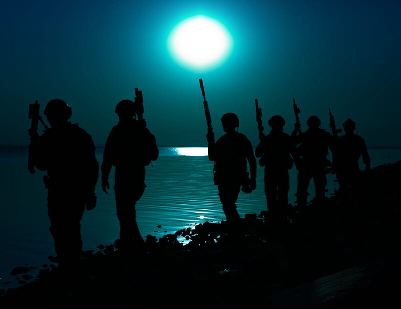 Silhouetted military service members carrying rifles walk along a shoreline at night