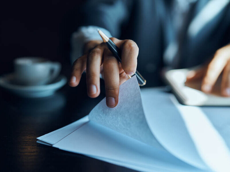 Man in a suit turning a page of a document stack on a dark desk