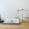 Wooden judge's gavel resting on its round sound block beside a set of evenly balanced brass scales of justice, with an open clipped document