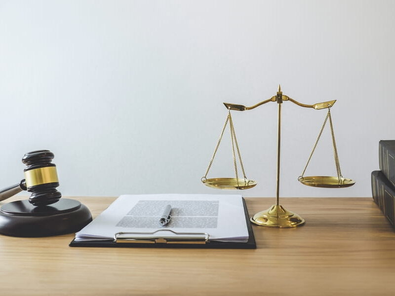 Wooden judge's gavel resting on its round sound block beside a set of evenly balanced brass scales of justice, with an open clipped document